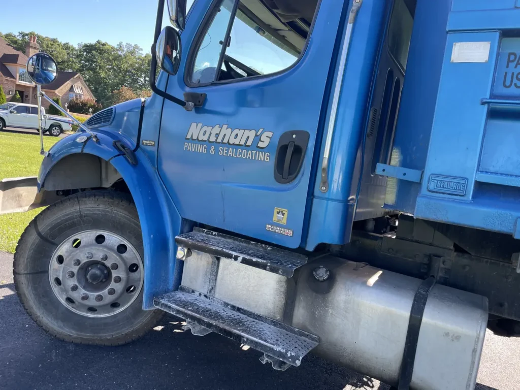 A close-up of the driver's side of a blue truck labeled "Nathan's Paving & Sealcoating," a premier driveway replacement company, parked on a paved road with grass and houses visible in the background.