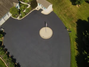 Aerial view of a circular driveway with a flagpole in the center, surrounded by well-manicured lawns and bushes, adjacent to a house and garage. Recent driveway sealing by Carlisle experts enhances the property's pristine appearance.