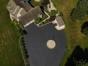 Aerial view of a property featuring a main house, a circular paved driveway with a central pole, expertly maintained through driveway sealing Carlisle, a smaller building, and surrounding landscaped greenery.