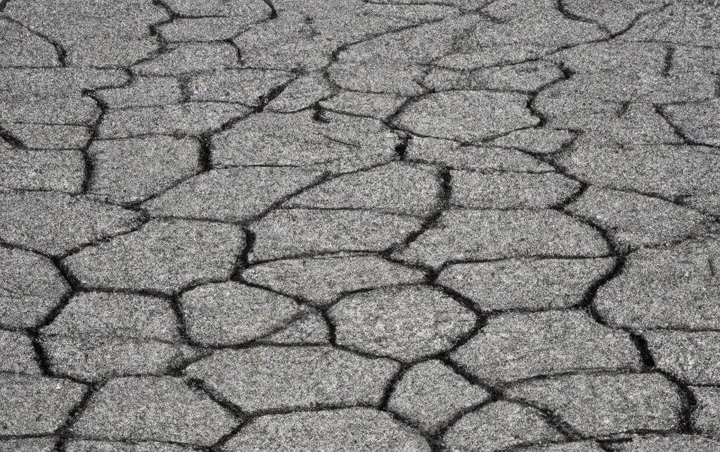 A close-up view of an asphalt driveway featuring alligator cracks, characterized by a network of interconnected fissures resembling the skin of an alligator. The surface is weathered and uneven.