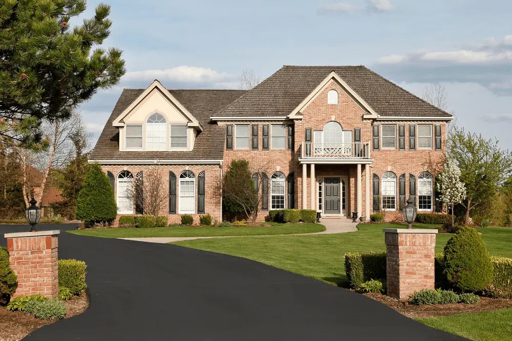 A large brick home with a blacktop driveway.|-up of a person wearing protective gloves and safety glasses spraying a pressure washer on a blacktop driveway|P of a person carefully rolling a thick layer of black asphalt sealant on a newly-laid driveway|-up of a squeegee tool being used to spread fresh blacktop on an asphalt driveway|Top driveway with an area freshly patched