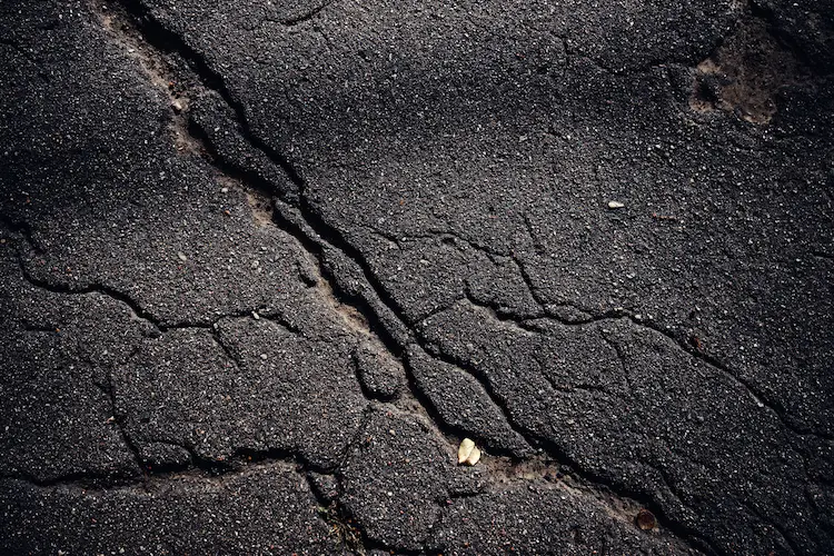 Cracks on a black asphalt driveway that require crack repair before sealcoating.|A road with cracks in it at night.|Cracks in the road with a person walking in front of a car