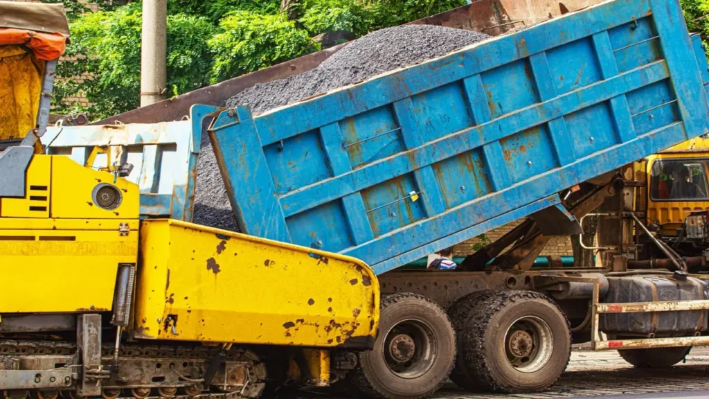 A freshly laid asphalt surface being loaded into a large construction truck at a paving site. The asphalt is dark and smooth, indicating recent application and preparation for roadwork.