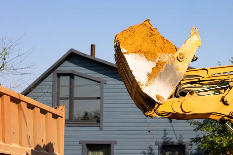 Close-up of a construction excavator's bucket pouring sand into a container, with a residential house in the background under clear skies