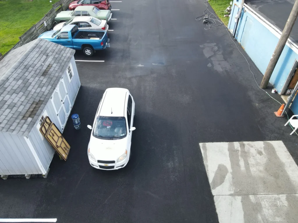 A newly paved parking lot features smooth asphalt with clear, white parking stripes. Freshly marked spaces are visible, and the surrounding area includes grass and a few trees in the background.