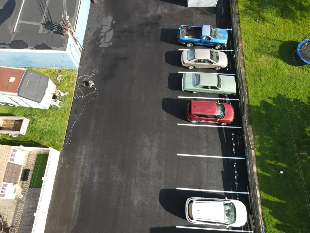 An aerial view of a freshly paved parking lot with crisp, clean lines. The surface is smooth and dark asphalt, surrounded by green grass and a blue sky in the background.