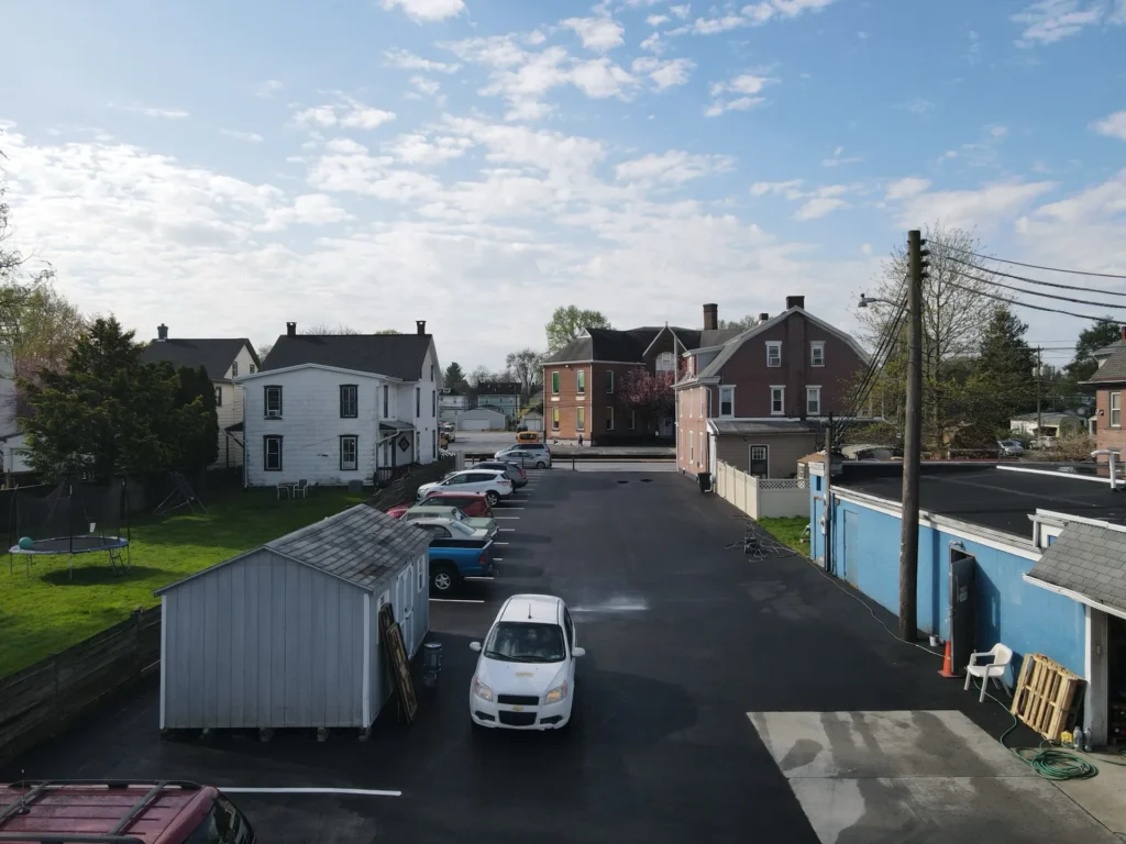 A newly paved parking lot with a smooth black surface, marked parking spaces, and clear white lines. Surrounding greenery and a clear blue sky complement the scene.