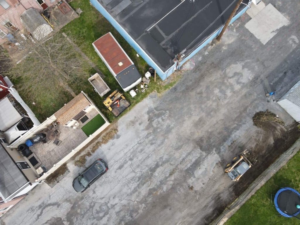 An asphalt parking lot in poor condition, with visible cracks and patches. The surrounding area features greenery and a clear blue sky. The image is framed to highlight the lot's surface wear.