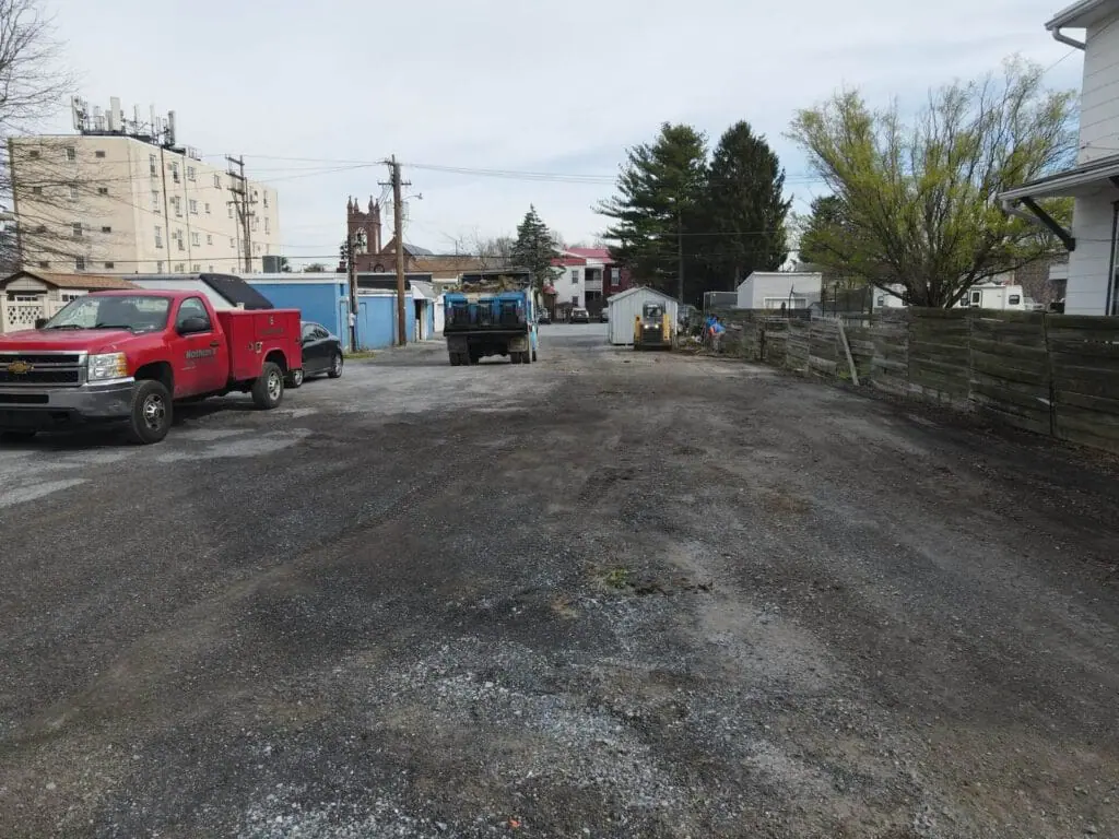 Alt text: A parking lot before paving, featuring uneven ground with visible dirt, gravel, and sparse vegetation. There are several parked cars, indicating prior use of the area.