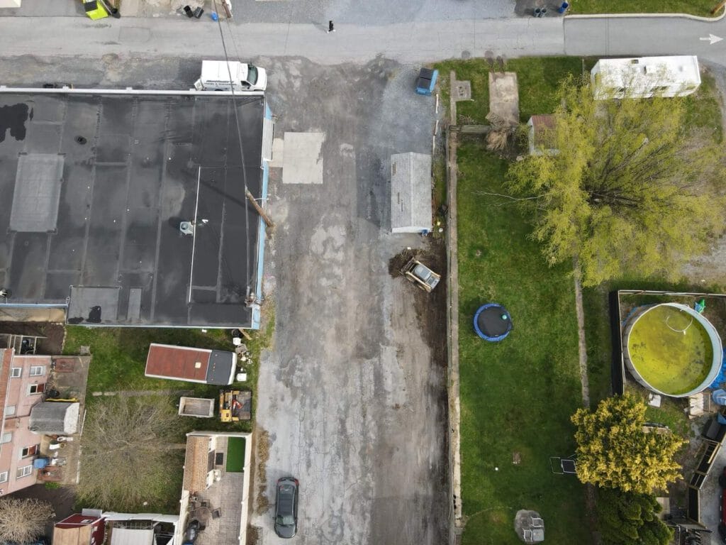 A spacious parking lot before asphalt paving, featuring uneven gravel, scattered debris, and unclear road markings under a cloudy sky. Surrounding greenery is visible in the background.