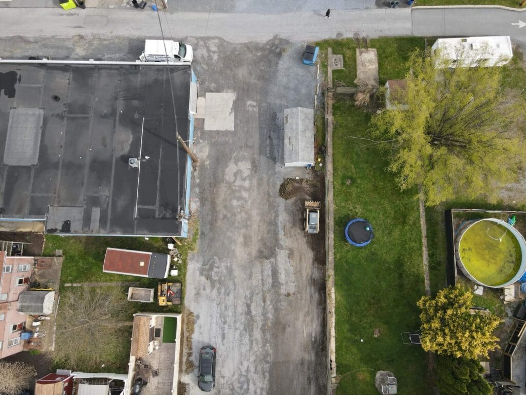 An aerial view of a parking lot before renovation, featuring uneven asphalt surfaces, cracks, and weeds. Surrounding trees and a building in the background indicate a commercial area.