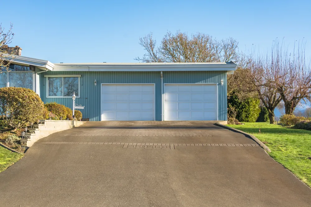 A home with two garages and a driveway that meets width requirements.|Two cars parked in a driveway.|A person's hand holding a measuring tape to determine driveway width requirements on a street.