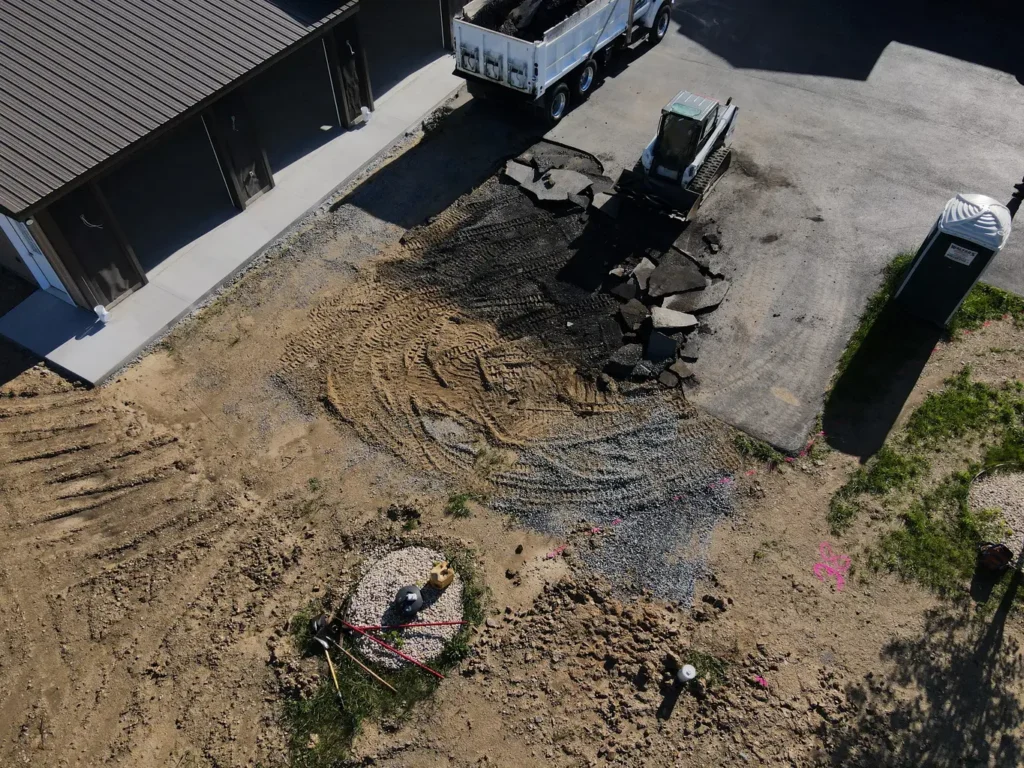 A before photo of a driveway extension project in Carlisle, featuring an unpaved, uneven area of gravel and grass adjacent to a paved driveway, indicating the space for future expansion.