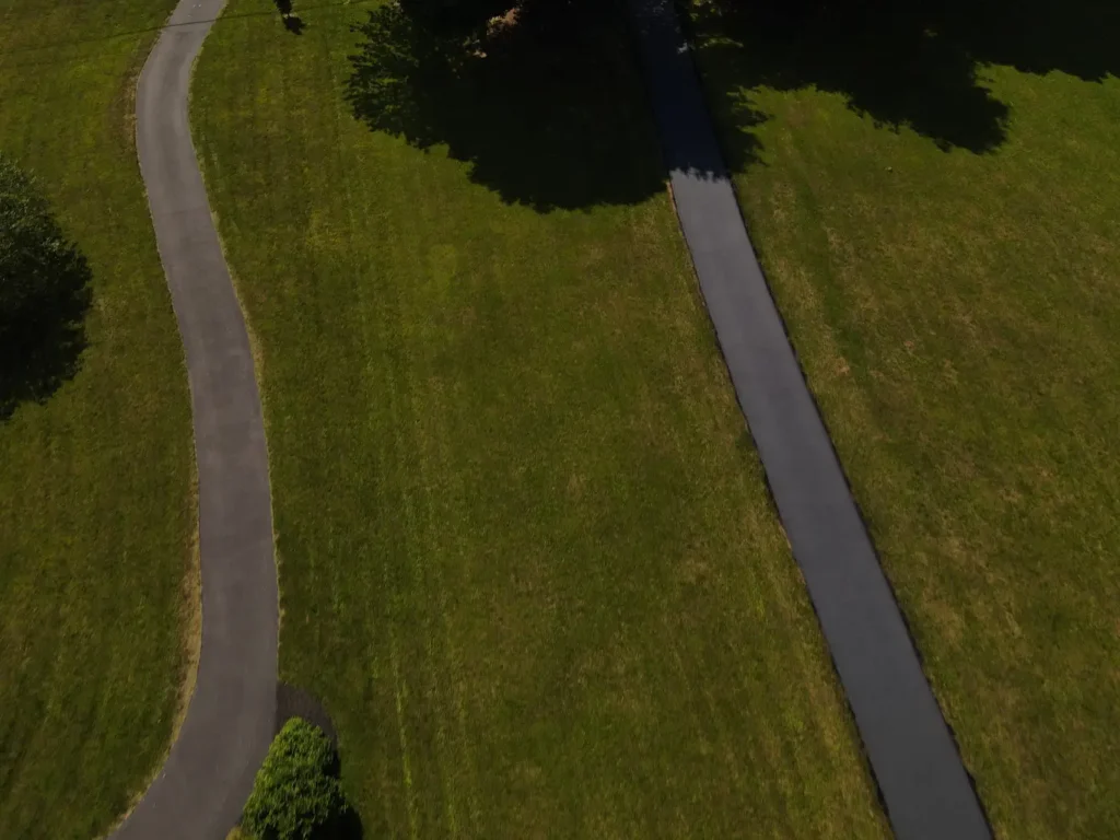 A freshly sealed black asphalt driveway, showing a smooth, shiny surface with a slight shine under sunlight. Surrounding grass is green, and the driveway’s edges are well-defined.