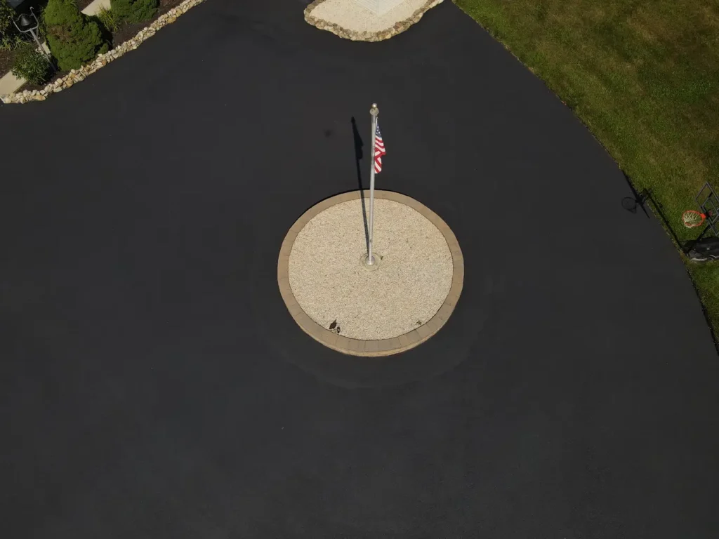 A freshly sealed black asphalt driveway is shown, showcasing a smooth and glossy surface. The driveway is bordered by grass and trees, indicating a residential setting in Carlisle.