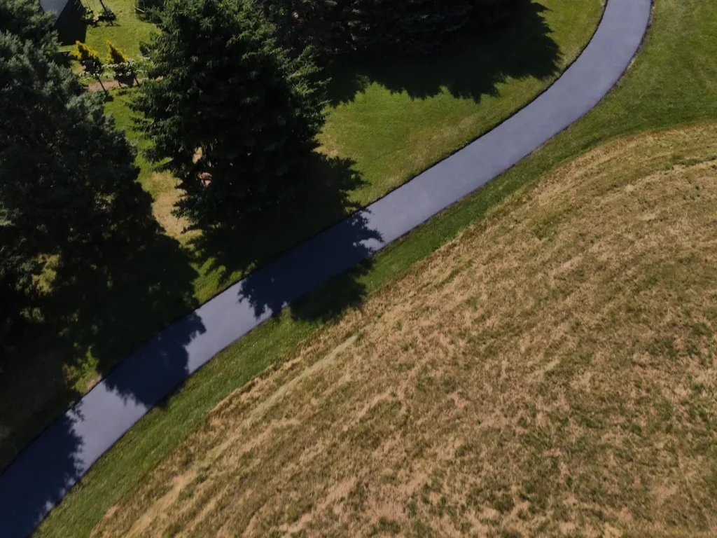 The image shows a freshly sealed asphalt driveway in Carlisle, featuring a smooth, shiny black surface under clear blue skies and a line of green grass along the border.