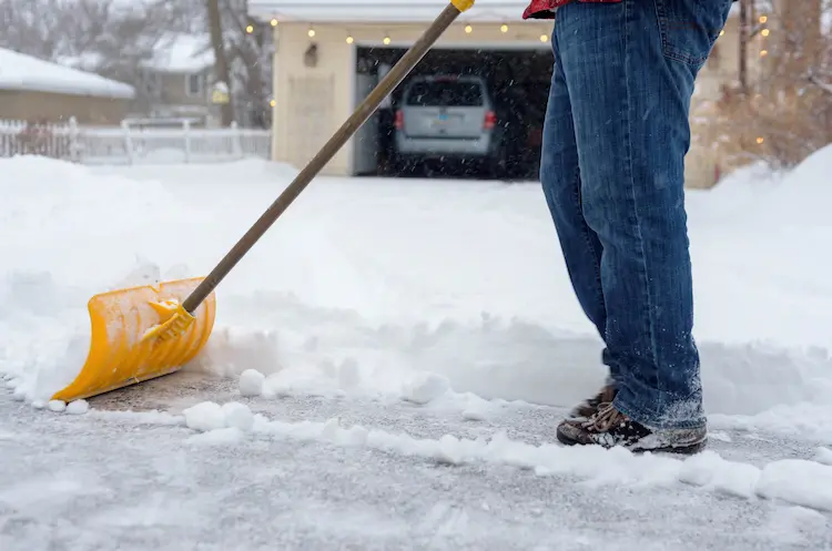 A man with a yellow shovel is clearing snow from a driveway, ensuring the proper maintenance of the asphalt paving.