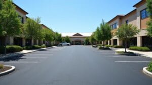A wide, empty parking lot in Camp Hill, bordered by two rows of trees and office buildings under a clear blue sky. The pristine condition reflects meticulous parking lot maintenance ensured by local experts.