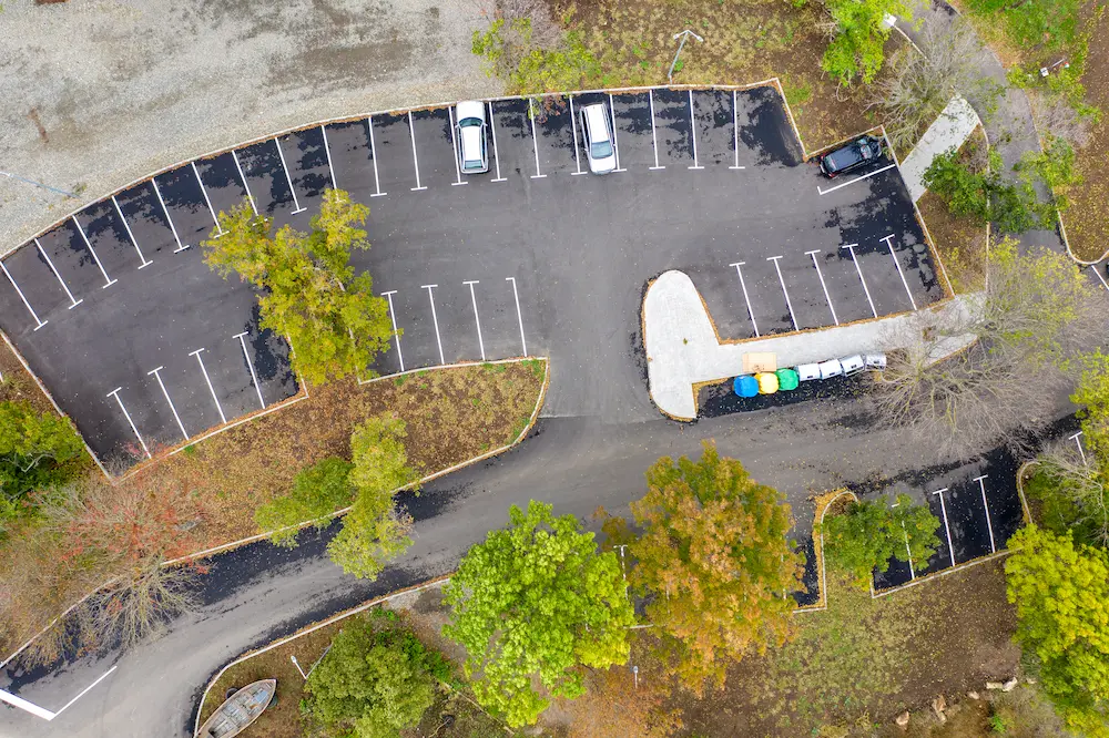 Aerial view of a small, mostly empty parking lot with a few parked cars. Several trees and a surrounding grassy area are visible.